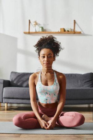 Mujer afroamericana rizada en ropa deportiva haciendo yoga en una estera en un acogedor entorno de sala de estar.