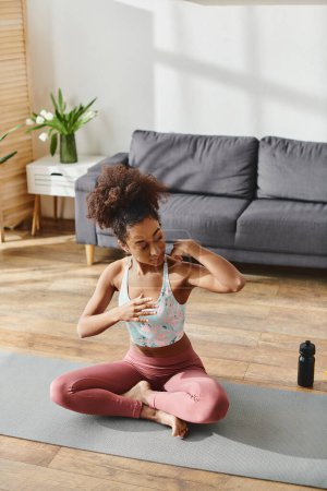 A curly African American woman in activewear practices yoga on a mat in a cozy living room setting.