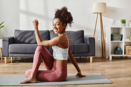 Femme afro-américaine bouclée en tenue active pratiquant le yoga sur un tapis dans un cadre confortable salon.