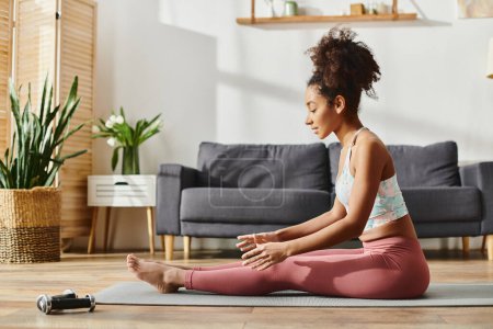 Femme afro-américaine frisée en tenue active pratique le yoga sur un tapis dans un salon confortable.