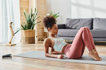 Femme afro-américaine bouclée en tenue active exécute gracieusement une pose de yoga sur un tapis de yoga coloré à la maison.