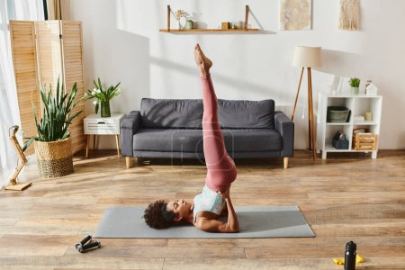 Mujer afroamericana rizada en ropa deportiva que muestra equilibrio y fuerza haciendo un soporte de mano en una esterilla de yoga en casa.