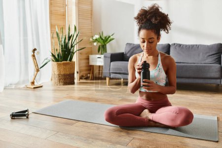 Lockige Afroamerikanerin in Aktivkleidung sitzt auf einer Yogamatte und hält friedlich eine Flasche Wasser in der Hand.