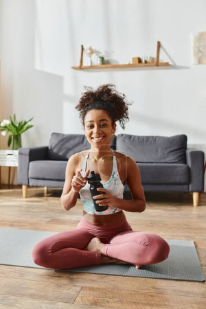 Una mujer afroamericana rizada en ropa deportiva se sienta en una esterilla de yoga, sosteniendo una botella de agua.