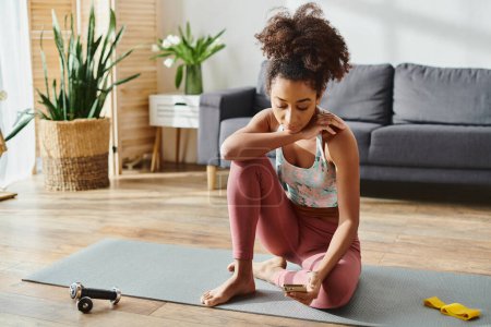 Foto de Una mujer afroamericana rizada en ropa activa sentada en una esterilla de yoga en una acogedora sala de estar, practicando yoga posa. - Imagen libre de derechos