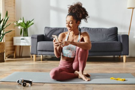 Lockige Afroamerikanerin in Activwear sitzt auf Yogamatte, in Telefonbildschirm vertieft.