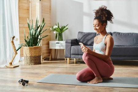 African American woman in activewear, sitting on yoga mat, focused on phone screen.