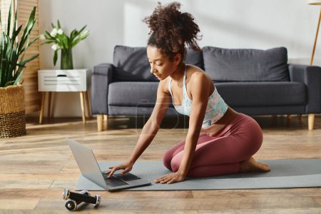 Foto de Mujer afroamericana rizada en ropa deportiva equilibra en una estera de yoga mientras usa una computadora portátil. - Imagen libre de derechos