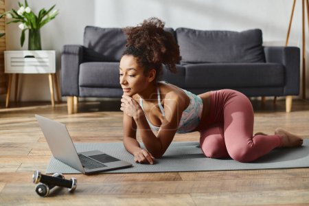 Una mujer afroamericana rizada en ropa deportiva usa una computadora portátil mientras está acostada en una esterilla de yoga en casa.
