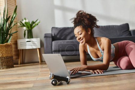 Una mujer afroamericana rizada en ropa activa, tendida en el suelo, se centra en su computadora portátil mientras hace ejercicio en casa.