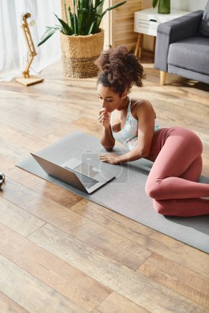Foto de Una mujer afroamericana rizada en uso activo practica yoga en una estera mientras usa una computadora portátil en casa. - Imagen libre de derechos