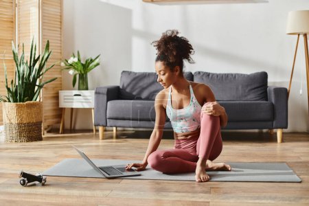 Foto de Una mujer afroamericana rizada en activo se viste pacíficamente sentada en una esterilla de yoga en una acogedora sala de estar. - Imagen libre de derechos