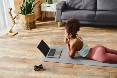 Foto de Mujer afroamericana rizada en ropa deportiva sentada en una esterilla de yoga, utilizando una computadora portátil para la instrucción virtual de fitness. - Imagen libre de derechos