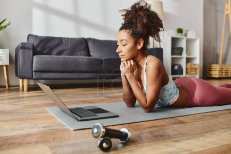 Una mujer afroamericana rizada en ropa deportiva practica yoga mientras usa una computadora portátil en una alfombra en casa.