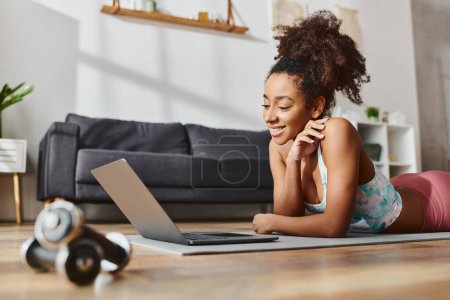 Una mujer afroamericana rizada en ropa activa que trabaja en casa, usando una computadora portátil mientras está acostada en el suelo.