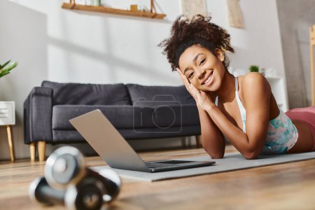 Une femme afro-américaine active dans les types de vêtements d'entraînement sur un ordinateur portable tout en étant couché sur le sol.
