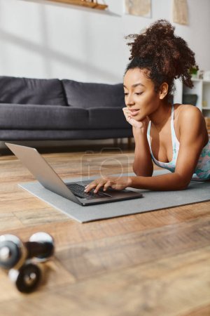 Curly African American woman in active wear uses laptop, laying on yoga mat, blending technology with physical activity.