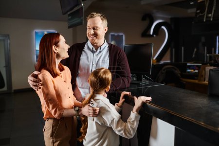 A man and woman stand next to a little girl, cherishing a day out together as a happy family at the cinema.