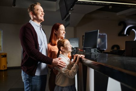 Un homme, une femme et un enfant heureux debout à un comptoir de concession dans un cinéma, choisissant des collations avant de profiter d'un film ensemble.