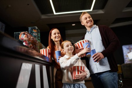 A man, woman, and child cheerfully hold popcorn boxes while spending quality time together at the cinema.
