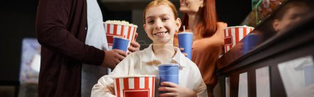A young girl joyfully holds bucket of popcorn at the cinema with her family.