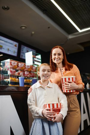 A woman stands next to a daughter, holding two boxes of popcorn, while enjoying a family trip to the cinema.