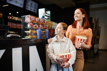 A happy woman stands next to a girl holding two buckets of popcorn at the cinema.