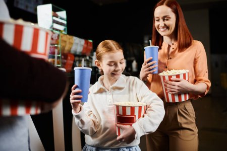 A woman and a girl happily holding cups of popcorn while spending family time at the cinema.