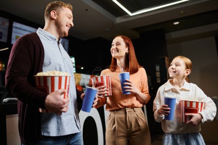 A happy family stands close, holding cups in a cinema, sharing a moment of togetherness and joy.
