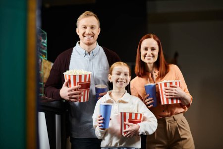 Téléchargez les photos : A man, woman, and child joyfully hold popcorn boxes at the cinema, bonding over snacks and quality time together. - en image libre de droit