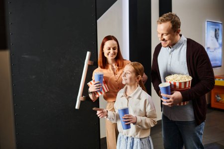 Un homme, une femme et un enfant tenant joyeusement des seaux de maïs soufflé au cinéma, profitant d'une sortie de cinéma en famille.