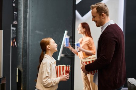 A man stands next to a little daughter, both happily holding popcorn at the cinema.