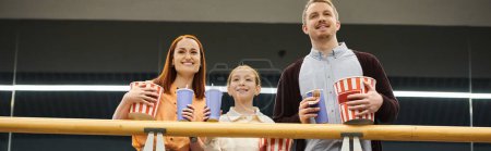 A happy family standing together, holding cups, enjoying a movie night at the cinema.