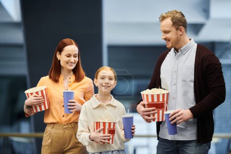 A man, woman, and child happily hold boxes of popcorn while spending time together in a cinema.