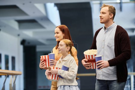 A man, woman, and child happily hold popcorn boxes during a fun family movie night at the cinema.