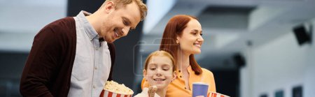 Un hombre está al lado de una esposa y un niño, ambos sonriendo, mientras ella sostiene una caja de palomitas de maíz en un cine.