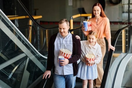 Téléchargez les photos : Un homme et deux petites filles descendent joyeusement un escalier roulant ensemble dans un cinéma, créant une scène de famille chaleureuse. - en image libre de droit