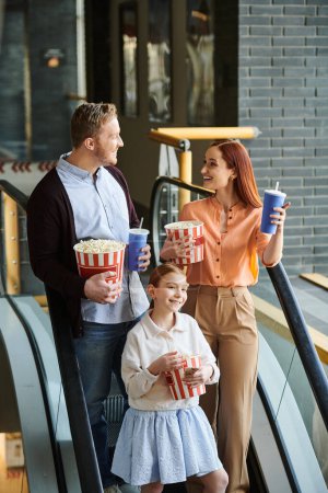 Téléchargez les photos : Une famille joyeuse de trois personnes se tient joyeusement sur un escalier roulant dans un cinéma, profitant de leur temps ensemble. - en image libre de droit