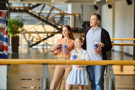 Un homme, une femme et un enfant tiennent joyeusement des tasses et du pop-corn, profitant d'un moment ensemble dans un cinéma.