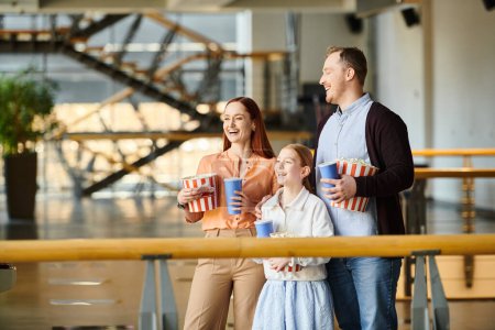 A happy family stands close, each holding a cup as they enjoy a movie together in the cinema.