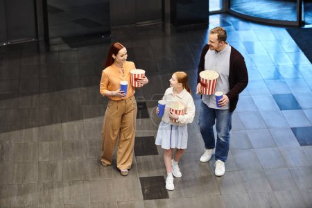 father and son hold popcorn while a little girl stands next to them at the cinema, enjoying a happy family moment.