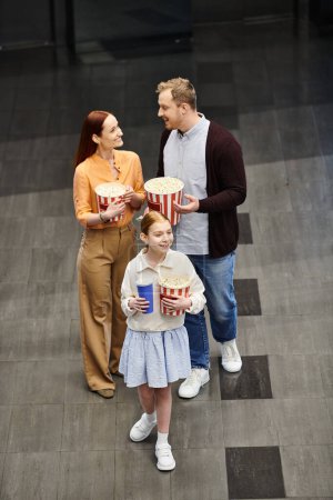 family happily hold popcorn buckets while standing next to a little girl, enjoying a movie night at the cinema.