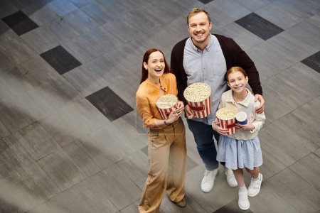 A man and his family happily hold popcorn boxes at the cinema, enjoying a family movie night together.