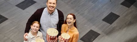 A man, woman and girl happily holding popcorn boxes at the cinema.