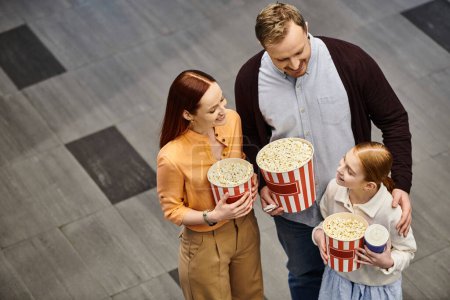 A man and his wife joyfully hold popcorn boxes with kid as they enjoy a movie together at the cinema.