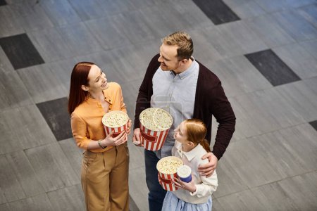 A man and woman excitedly hold popcorn buckets while a child smiles, creating a happy family scene at the cinema.