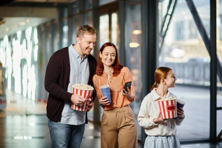 Un couple tenant joyeusement un seau de pop-corn près de sa fille, profitant de moments de qualité ensemble au cinéma.