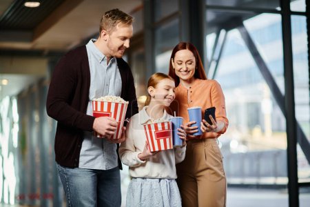 A man, woman, and child happily hold popcorn boxes while enjoying a movie together at the cinema.