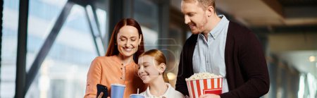 A happy family enjoys popcorn together at the cinema, standing around a table with a variety of snacks.