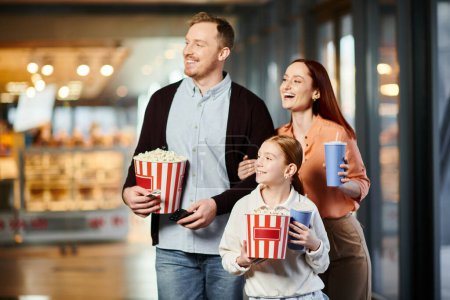 A man, woman, and child happily hold popcorn and sodas while spending quality time together at the cinema.